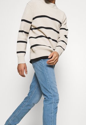 Beige geribbelde trui met zwarte horizontale strepen, gedragen met lichtblauwe jeans. De trui heeft een hoge halslijn en aansluitende manchetten.