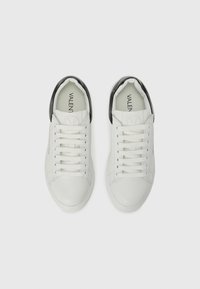 Hvite skinn-sneakers med en svart hælflik, rund tå og flate snører. Glatt tekstur med minimal merkevare på tungen.