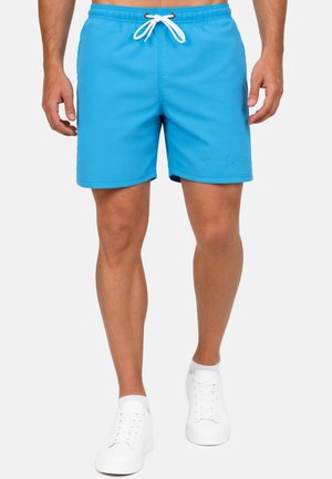 Uomo che indossa pantaloncini da bagno blu brillante con coulisse bianca e sneakers bianche, in piedi davanti a uno sfondo semplice.