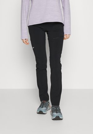 Person iført sorte, smalle bukser med sidelomme, lyse lilla langærmet top og grå vandresko, der står på gråt gulv.