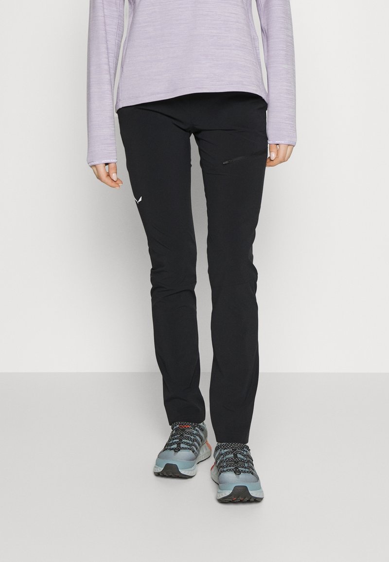 Personne portant un pantalon noir slim avec une poche latérale, un haut violet clair à manches longues et des chaussures de randonnée grises, debout sur un sol gris.