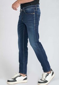 Jeans en denim bleu foncé avec une coupe slim, présentant un léger délavage, cinq poches et des coutures contrastées, portés avec des baskets noires et blanches.