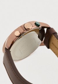 Close-up of a rose gold watch with brown leather strap, showing side buttons and engraved back with brand and material details.