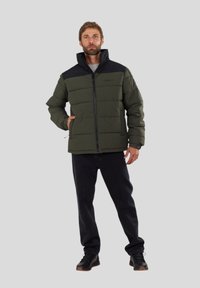 Green puffer jacket with black accents, high collar, front zip, and horizontal quilting; paired with dark pants and black shoes.