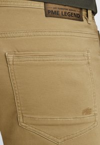 Beige pants featuring a classic cut, with a back pocket, stitched detailing, and a logo patch at the waistband. Smooth fabric texture.