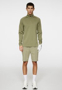 Olivgrön långärmad zip-up-tröja, ljusgröna shorts, vita strumpor, vita sneakers med svarta detaljer och en vit handske.