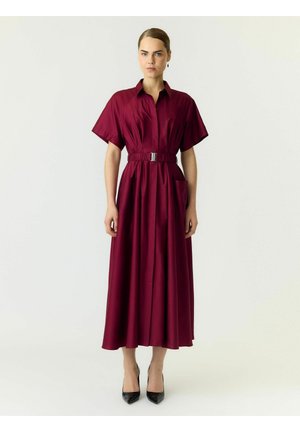 Kvinde stående iført en burgundy midi kjole med bælte og korte ærmer og sorte spidse hæle på en ensfarvet lys baggrund.