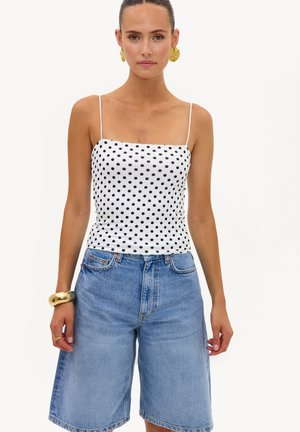 Femme portant un débardeur blanc à fines bretelles avec des pois noirs et un short en jean bleu ample, agrémentée de boucles d’oreilles et d’un bracelet dorés.