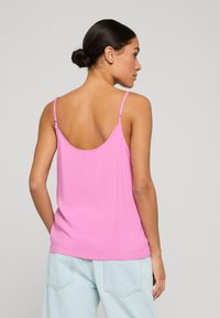 Mujer con cabello oscuro recogido en un moño, que lleva una blusa sin mangas rosa y vaqueros azul claro, de espaldas sobre un fondo gris liso.