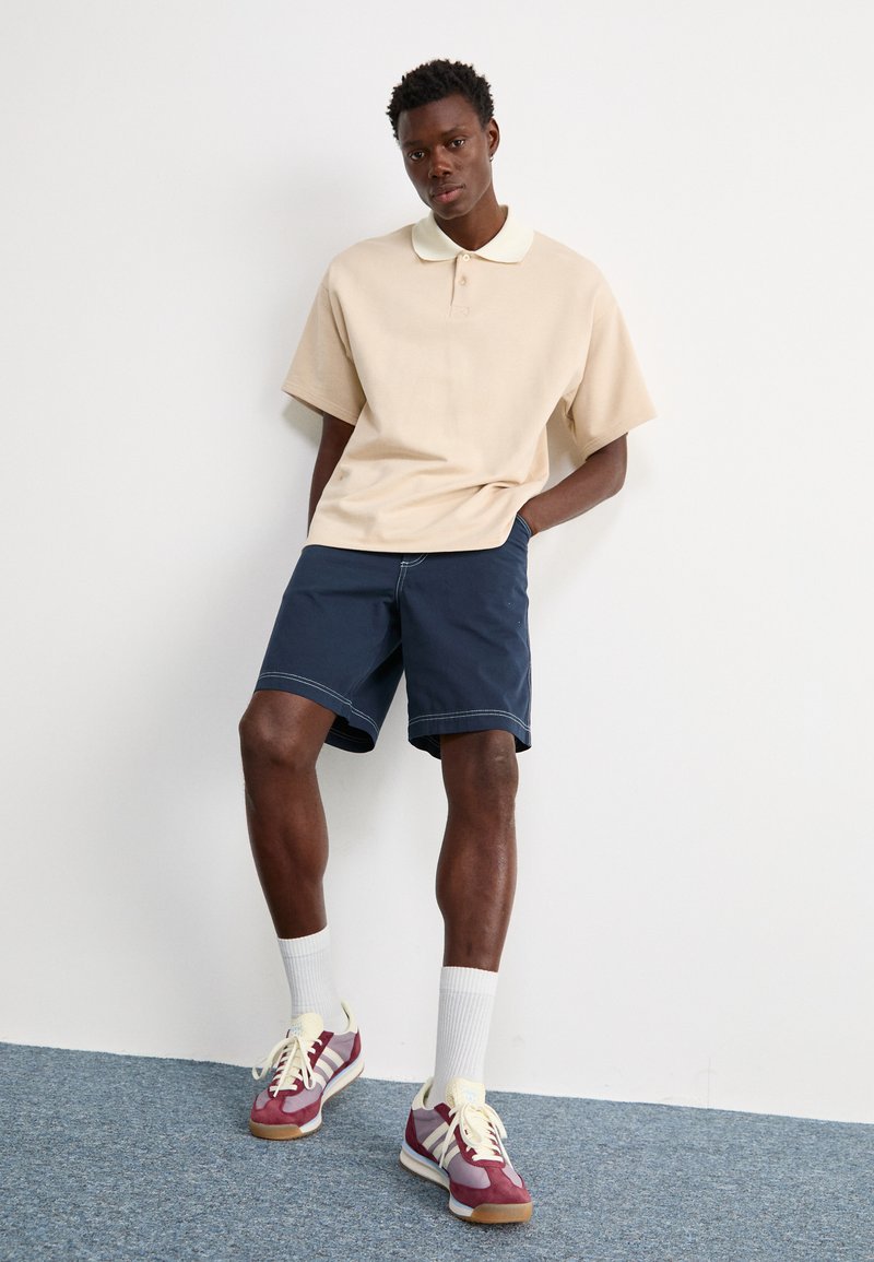 Polo beige avec un col blanc, associé à un short bleu marine. Le modèle porte des chaussettes blanches et des baskets multicolores avec des accents.