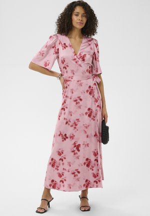 Kobieta w różowej sukience kopertowej w kwiaty i czarnych sandałach, trzymająca czarną plecioną kopertówkę, stojąca z jedną ręką na biodrze.