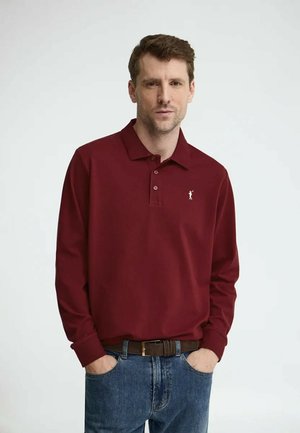 Uomo con capelli castani corti, indossa una polo a maniche lunghe color borgogna e jeans blu, in piedi con le mani in tasca davanti a uno sfondo semplice.