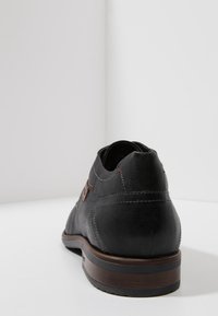 Chaussure en cuir noir à texture lisse, talon arrondi, accents en bois marron et détail de couture rouge sur la tige.