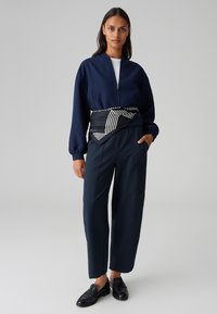 Veste de bombardier marine avec fermeture éclair, associée à un pantalon navy à coupe ample. Une ceinture noire et blanche rayée souligne la taille. Des mocassins noirs complètent le look.