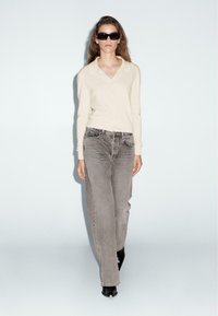 Beige cardigan with a collared design, paired with loose gray jeans and black heeled boots. Model wears black sunglasses. Neutral background.