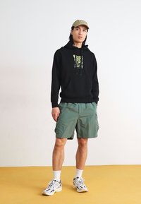 Black hoodie with a graphic print, green cargo shorts, white sneakers with black accents, and a light green cap.