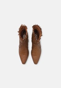 Botas de tobillo trenzadas marrón con punteras puntiagudas, bordes con flecos y cremalleras laterales, hechas de un material de cuero texturizado.