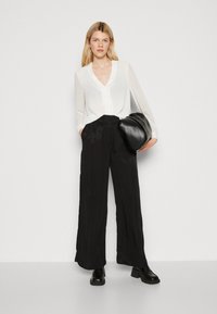 Blusa bianca a maniche lunghe con scollo a V, abbinata a pantaloni neri a gamba larga con un motivo testurizzato. Borsa nera e scarpe con tacco chunky.