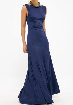 Kobieta w granatowej, bezrękawowej sukni sięgającej podłogi, z drapowanym materiałem i masywnym naszyjnikiem z koralików na białym tle.