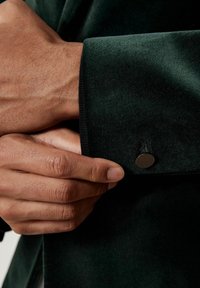 Tume roheline sametist särgi varrukas, millel on sile pinnatunne, kinnitatud ümmarguse musta nööbiga. Nähtav käsi haarab varrukast.