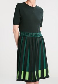 Robe vert foncé à manches courtes, avec une jupe plissée et une ceinture verte à motif. Détails d'ourlet en vert clair en accent.