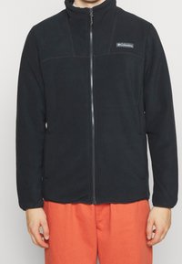 Giacca in pile nera con colletto alto, cerniera completa e logo sulla parte superiore sinistra. Presenta una superficie liscia e due tasche laterali.