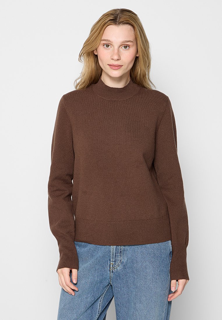 Brauner Strickpullover mit hohem Kragen, langen Ärmeln sowie gerippten Bündchen und Saum. Kombiniert mit hellblauen Jeans.