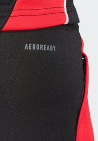 Zwarte sportstof met rode accenten, voorzien van een ritszak en "AEROREADY" branding. De textuur is glad met een vochtregulerend ontwerp.