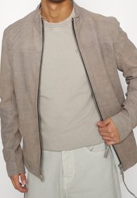 Svetle šedá semišová bunda s stojacími límcem, předním zipem a tónovaným prošíváním. Nasažená přes světlou béžovou košili s krátkými rukávy.