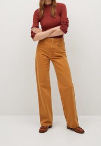 Nainen seisoo käsivarret ristissä, päällään ruosteenvärinen ribbipusero, korkea vyötärö ja leveälahkeiset sinapinkeltaiset housut sekä ruskeat frilleillä varustetut loafer-kengät.