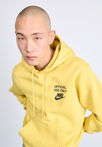 Sárga kapucnis pulóver fekete "HIVATALOS HASZNÁLATRA" felirattal és Nike logóval. Kenguru zsebbel és állítható kapucnival rendelkezik. Simább anyagú textúra.