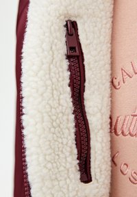 Burgunderfarvet jakke med en cremet fleece-foring og lynlås. Den strukturerede stofkontrast med den glatte lynlåsdesign. Lys pink broderet detalje synlig.