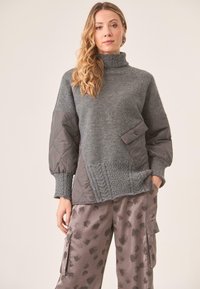 Suéter gris esponjoso con cuello alto, detalles en punto texturizado y mangas acolchadas. Combinado con pantalones satinados grises con un patrón de hojas.