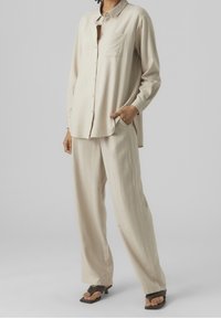 Beige langärmliges Hemd mit Knopfleiste und Tasche, kombiniert mit lockeren beigen Hosen, beide aus leichtem Material. Flache Sandalen.