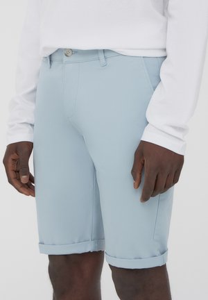 Pantaloni corti azzurri chiari con una texture liscia, chiusura con bottone e risvolti, indossati con una camicia bianca a maniche lunghe.