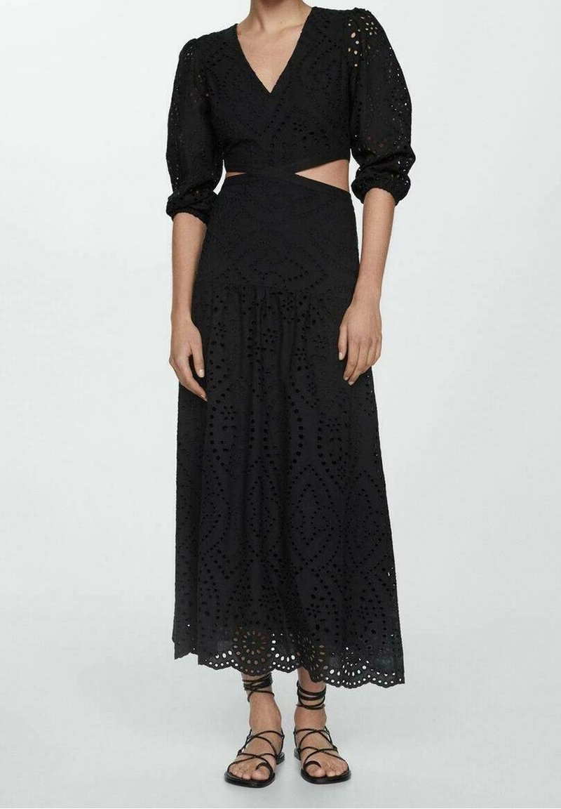 Robe maxi noire avec découpe à la taille, manches bouffantes et motif floral en œillets. Comprend une jupe évasée et un ourlet festonné. Associée à des sandales noires.