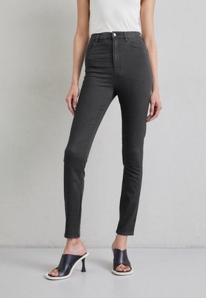 Frau trägt schwarze High-Waist Skinny-Jeans und schwarze offene Sandaletten mit Absatz, steht vor einer schlichten hellgrauen Wand.