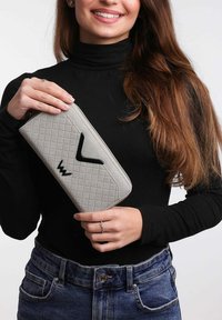 Šedá prošívaná peněženka s černými akcenty, segeometrickým designem, držena v rukou, na neutrálním pozadí.