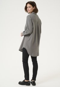 Langärmliges, graues Hemd mit abgerundetem Saum, Kragen und Rückenpasse. Kombiniert mit engen schwarzen Jeans und schwarzen Slippern.