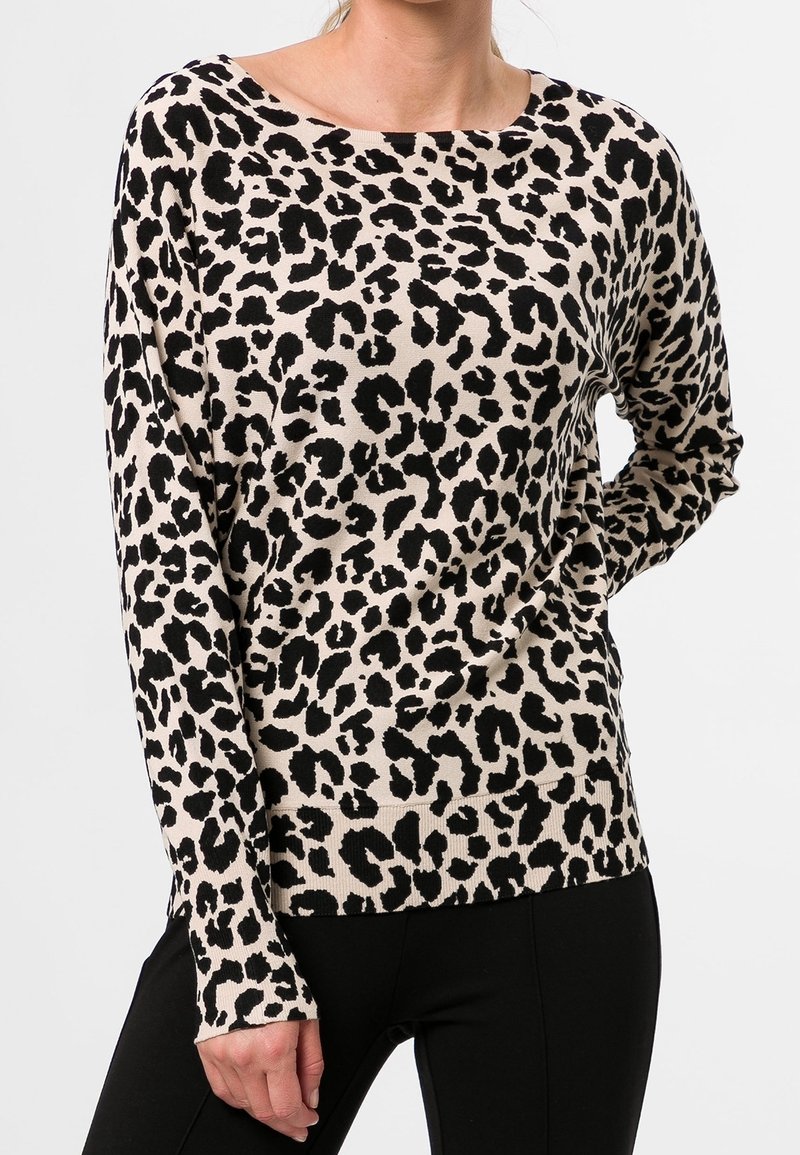 Femme portant un pull à manches longues beige imprimé léopard noir et un pantalon noir, debout avec une main sur la hanche.