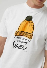 Camiseta de algodón blanca con un gorro de invierno amarillo impreso que tiene un pompón verde y detalles en línea negra. El texto dice "Altonadock company Winter #25".