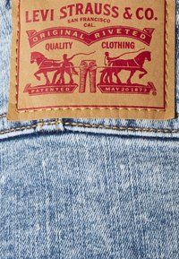 Toppa Levi Strauss & Co. su denim; pelle marrone con grafiche stampate in rosso, che raffigurano cavalli e jeans, cucita sul bordo.
