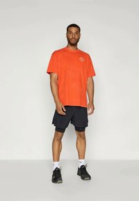 Oranges Baumwoll-T-Shirt mit dem Aufdruck "Nike Athletic Dept." , kombiniert mit schwarzen Shorts und schwarzen Sportschuhen. Einfaches Design, lässige Passform.