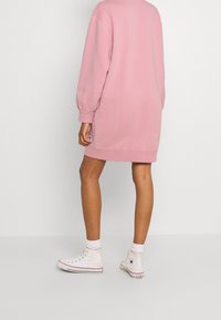 Rosa sweatshirtklänning med långa ärmar och avslappnad passform, tillverkad av ett mjukt tyg. Matchas med vita högskor och strumpor.