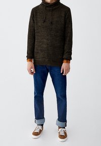 Pull en tricot vert foncé avec un col montant et des cordons de serrage, associé à un jean bleu retroussé aux chevilles et des baskets marron à semelles blanches.