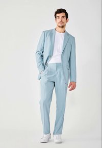 Completo azzurro chiaro con giacca monopetto, pantaloni coordinati e sneaker bianche. Texture liscia, design su misura e dettagli minimi.