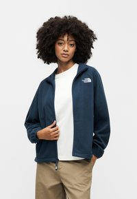 Σκούρο μπλε φλις μπουφάν με φερμουάρ, ψηλό γιακά και λογότυπο στο αριστερό μέρος του στήθους. Συνδυάζεται με λευκό πουκάμισο και μπεζ παντελόνι. Μαλακή υφή.