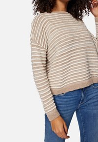 Beżowy sweter w paski o luźnym kroju, lekka dzianinowa faktura, szeroki dekolt i ściągacze na rękawach. Noszony z niebieskimi dżinsami.