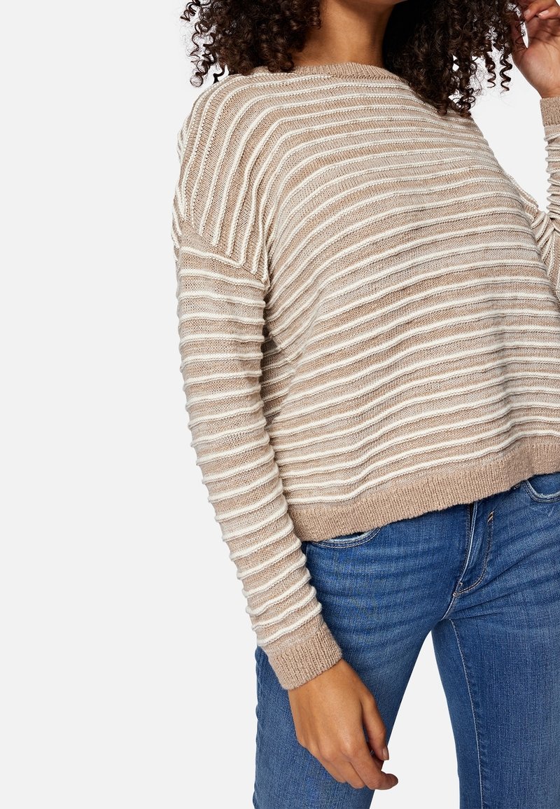 Beżowy sweter w paski o luźnym kroju, lekka dzianinowa faktura, szeroki dekolt i ściągacze na rękawach. Noszony z niebieskimi dżinsami.