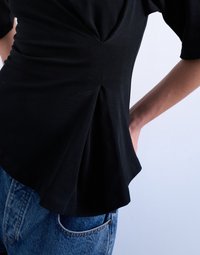 Μαύρο εφαρμοστό τοπ με λεπτομέρεια drape και ράβδωμα στη μέση, συνδυασμένο με μπλε τζιν, με το ένα χέρι να στηρίζεται στον γοφό.